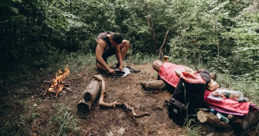 Backpacker preparing food at a forest campsite with a pack and camping equipment.